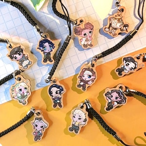 May include: A selection of wooden charm keychains with cartoon character designs. Each charm is attached to a black braided cord with a loop. The charms are in various colours and depict different characters.