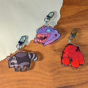 May include: Three colourful keychains featuring cartoon characters. The first keychain is a purple and blue monster with a toothy grin. The second keychain is a grey raccoon with black stripes. The third keychain is a red monster with a toothy grin.
