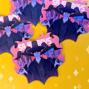 May include: A collection of stickers featuring a stylized illustration of a group of anime-style characters. The characters are depicted in dark clothing and hats, with glowing yellow eyes and purple skin. The stickers have a pink flame-like border with the text "SA IA BOYS".