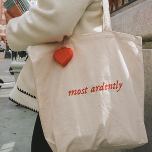 May include: A cream-colored tote bag with the words "most ardently" in red script. A small red heart charm hangs from the bag. The bag is carried on a shoulder. The background shows a city street with buildings and a traffic cone.