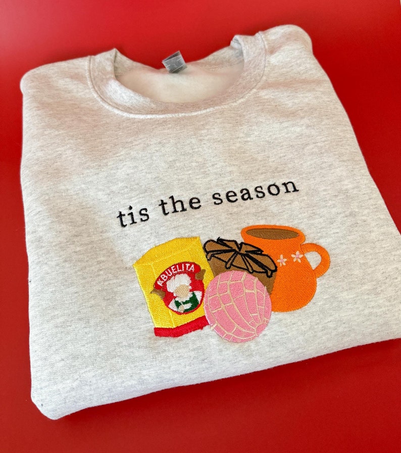 Puede incluir: Sudadera gris con el texto "tis the season" bordado en la parte delantera. El texto est&aacute; rodeado de im&aacute;genes bordadas de un pan dulce, una taza de chocolate caliente y una caja de chocolate caliente Abuelita.