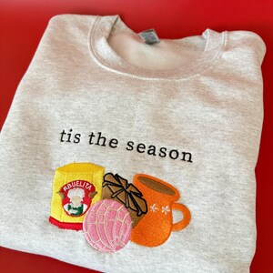 Puede incluir: Una sudadera gris con el texto "tis the season" bordado en la parte delantera. Debajo del texto hay una imagen bordada de una taza de caf&eacute;, un pan dulce, una barra de chocolate y una caja de mezcla de chocolate caliente Abuelita.