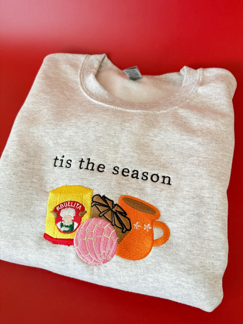 Puede incluir: Una sudadera gris con el texto "tis the season" bordado en la parte delantera. Debajo del texto hay una imagen bordada de una taza de caf&eacute;, un pan dulce, una barra de chocolate y una caja de mezcla de chocolate caliente Abuelita.