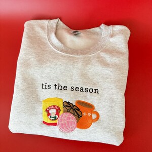 Puede incluir: Una sudadera gris con el texto "tis the season" bordado en la parte delantera. La sudadera presenta un dise&ntilde;o bordado de una taza de chocolate caliente, un pan dulce y una caja de chocolate caliente Abuelita.