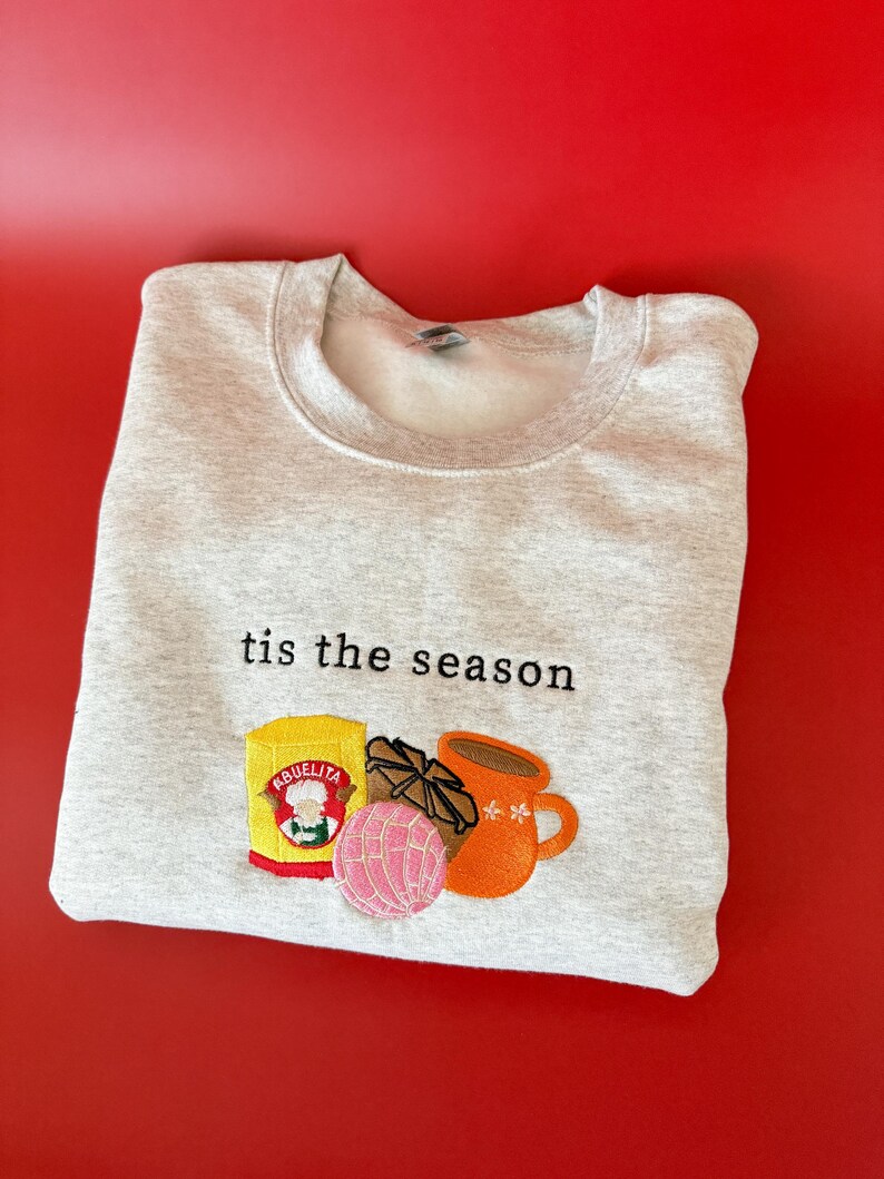 Puede incluir: Una sudadera gris con el texto "tis the season" bordado en la parte delantera. La sudadera presenta un dise&ntilde;o bordado de una taza de chocolate caliente, un pan dulce y una caja de chocolate caliente Abuelita.