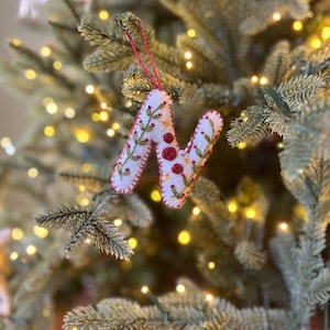 May include: White felt letter N ornament with red and green embroidery. The ornament is hanging on a Christmas tree with white lights.