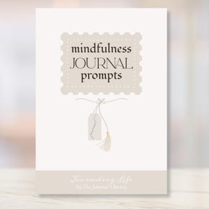 May include: A journal with the words "mindfulness JOURNAL prompts" in a decorative stamp-like design. Below the stamp is a small tag with the word "journal" and a tassel. The journal is cream-colored with the text "Journaling Life by The Journal Library" at the bottom.