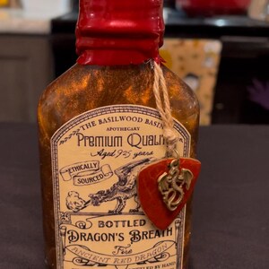 May include: A small, clear glass bottle with a red wax seal and a decorative tag. The label reads "Dragon's Breath Fire" and "Ancient Red Dragon." The bottle contains a shimmering, amber-colored liquid. A small charm with a dragon is attached.