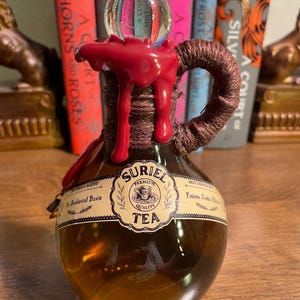 May include: A glass bottle with a red wax seal and a cork stopper. The bottle is labeled "SURIEL PREMIUM QUALITY TEA" and "The Blackwood Basin".