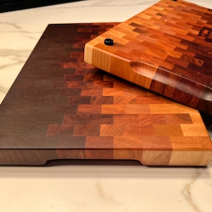 May include: Two wooden cutting boards with a gradient of brown, red, and light wood colors. The boards are made of small, square pieces of wood arranged in a mosaic pattern. The top board has a rounded edge and a handle.