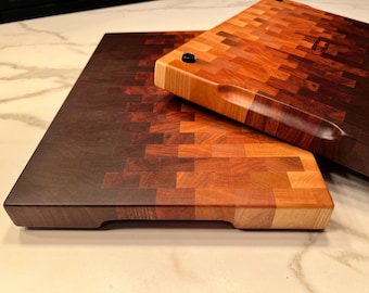 May include: Two wooden cutting boards with a gradient of brown, red, and light wood colors. The boards are made of small, square pieces of wood arranged in a mosaic pattern. The top board has a rounded edge and a handle.