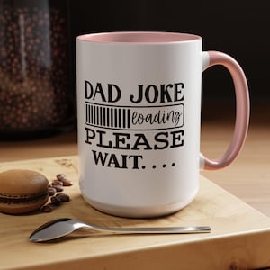 Op de afbeelding: Witte keramische mok met roze binnenkant en oor. De mok heeft de zwarte tekst "DAD JOKE loading PLEASE WAIT...". Een lepel, een macaron en koffiebonen liggen op een houten oppervlak.