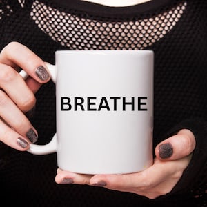 May include: White ceramic mug with the word "BREATHE" printed in black. The mug is held by a person wearing a black mesh top. The person's nails are painted with a glittery, dark color.