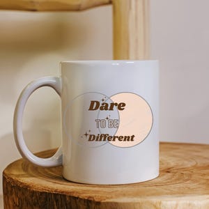 Może przedstawiać: Biały kubek ceramiczny z brązowym wzorem graficznym, na którym widnieje napis "Dare to be Different" z dwoma nakładającymi się na siebie okręgami i brzoskwiniowym tłem.
