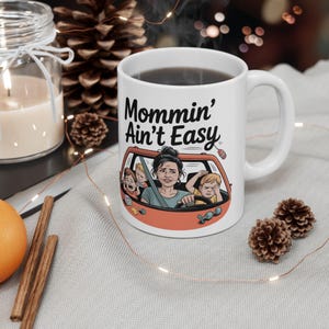 May include: White ceramic mug with the text "Mommin' Ain't Easy" and a cartoon illustration of a frazzled mother driving a car with three children. The mug is filled with a dark liquid.