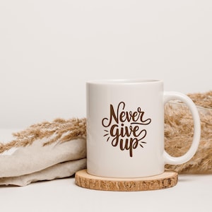 以下が含まれることがあります： 白いセラミック製のマグカップに、茶色の文字で「Never give up」と書かれています。マグカップは木製のコースターの上に置かれ、乾燥した草に囲まれています。