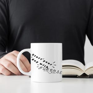 Puede incluir: Taza de café de cerámica blanca con un diseño negro que dice "Start your day with purpose".