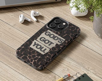 Op de afbeelding: Een zwarte telefoonhoes met een bruin en beige giraffenprintpatroon. De hoes heeft de tekst "GOD'S GOT YOU" in een verweerde, gebroken witte lettertype. De telefoonhoes ligt op een houten oppervlak met een laptop, oortelefoons en een potlood.