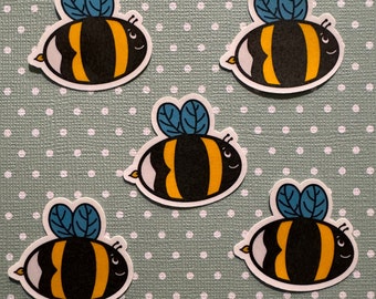 May include: Five cartoon bee stickers with black and yellow stripes, blue wings, and white eyes. The bees are on a light green background with white polka dots.