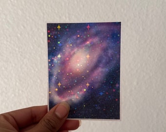 May include: A holographic sticker with a galaxy design. The galaxy is a swirl of pink, purple, and blue with a scattering of iridescent stars.