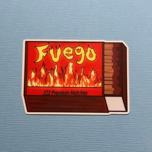 May include: A red and white matchbox with the word "FUEGO" in yellow letters and a graphic of flames. The box says "222 Premium Matches".
