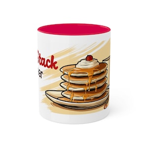 Puede incluir: Taza de cerámica blanca con borde rojo. La taza presenta una ilustración de dibujos animados de una pila de panqueques con jarabe y crema batida en un plato. El texto "Pancake Stack" está impreso en la taza.
