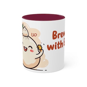 Könnte beinhalten: Weiße Keramiktasse mit einem burgunderfarbenen Rand. Die Tasse zeigt eine Cartoon-Illustration eines Bao-Brötchens mit einem lächelnden Gesicht und dem Text "Brewing with Bao" in Orange.
