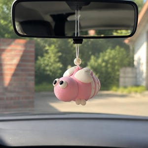 Könnte beinhalten: Ein rosa-weißes, bienenförmiges Auto-Ornament, das an einem Rückspiegel hängt. Die Biene hat einen abgerundeten rosa Körper mit weißen Flügeln, kleinen weißen Füßen und zwei großen schwarz-weißen Augen. Eine cremefarbene Perle sitzt über der Biene.