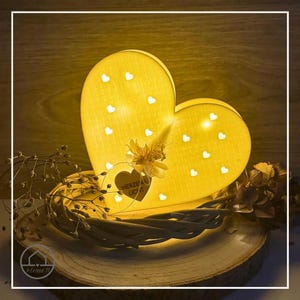 Könnte beinhalten: Eine herzförmige, gelbe Lampe mit kleinen Herz-Ausschnitten, von innen beleuchtet. Die Lampe ist mit Trockenblumen verziert und steht auf einem Holzsockel mit einem geflochtenen Kranz. Der Text "HERZEN LICHT" ist sichtbar.