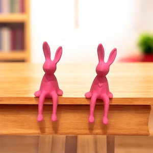 May include: Two bright pink bunny figurines sitting on the edge of a wooden table. The bunnies have long ears, small arms, and legs that dangle over the edge. One bunny has a heart on its chest. The background is blurred.