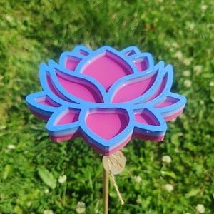 May include: A layered, three-dimensional lotus flower garden stake in shades of pink and blue. The flower is mounted on a wooden dowel with a natural twine accent. The stake is set in a bed of green grass.