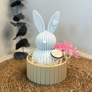 Könnte beinhalten: Hellblaue Osterhasenfigur mit Herzdetail am Ohr, in einem cremefarbenen Korb. Der Korb ist mit Stroh und rosa Blumen gefüllt. Ein Holzanhänger trägt die Aufschrift "Happy Easter".