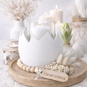 May include: A white egg-shaped candle holder with a jagged top, holding a lit white candle in a glass. The holder sits on a wooden tray with a wooden bead garland and a small wooden tag. Other candles and decorative elements are in the background.