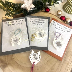 May include: Three small gift cards with white horseshoe charms, each with a different design inside. The cards have German text and are packaged in clear plastic. Festive decorations surround the cards.