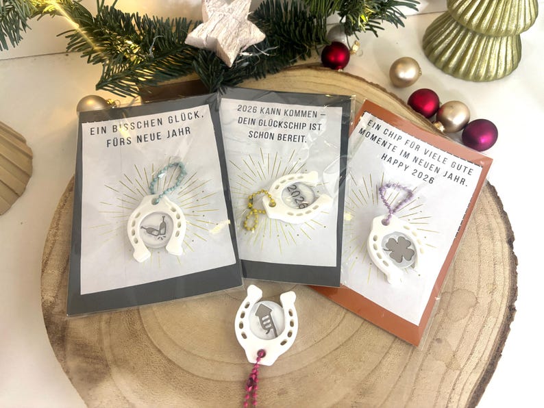 May include: Three small gift cards with white horseshoe charms, each with a different design inside. The cards have German text and are packaged in clear plastic. Festive decorations surround the cards.