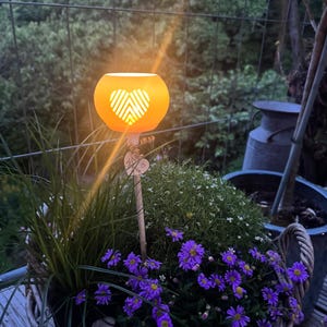 Puede incluir: Una lámpara solar de jardín naranja iluminada con un diseño de corazón, colocada en una cesta con flores moradas y vegetación. La lámpara está sobre una estaca de madera, añadiendo un toque decorativo.