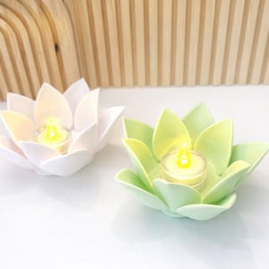 Könnte beinhalten: Zwei lotusblütenförmige Kerzenhalter, einer weiß und einer hellgrün, die jeweils ein kleines, brennendes Teelicht halten. Die Kerzenhalter stehen auf einer weißen Oberfläche, mit einem Holzbogen und einer Lamellenwand im Hintergrund.