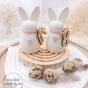 Può includere: Due statuette di conigli bianchi con dettagli a cuore ed etichette in legno, appoggiate su un vassoio di legno con uova macchiate e piume. I conigli hanno lunghe orecchie e sono decorati con spago. Il vassoio è su una superficie bianca.