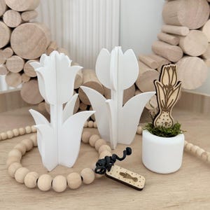 Op de afbeelding: Witte houten tulpdecoraties, waaronder twee grote 3D-tulpen en een kleinere houten tulp in een witte pot. Een houten kralenkrans en een klein houten label met een zwart touwtje zijn ook aanwezig.