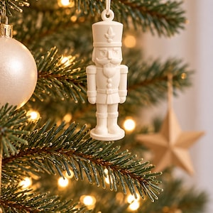 Könnte beinhalten: Ein mattweißer Nussknacker-Anhänger mit einem hohen Hut, der ein Fleur-de-Lis-Emblem trägt, sowie einem detaillierten Schnurrbart und einer Uniform mit Knöpfen. Die Figur hängt an einer dünnen weißen Schnur und ist einfarbig gestaltet.