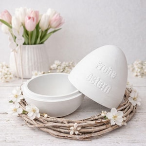 Könnte beinhalten: Ein weißer, eiförmiger Behälter mit den Worten "Frohe Ostern" auf dem Deckel, der auf einem Zweigkranz liegt. Der Behälter ist geöffnet. Eine Vase mit rosa und weißen Tulpen und eine Hasendekoration befinden sich im Hintergrund.