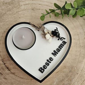 Puede incluir: Portavelas blanco en forma de corazón con borde negro. El portavelas tiene una pequeña hendidura redonda para una vela de té y las palabras "Beste Mama" están escritas en el portavelas.