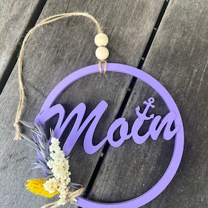 Puede incluir: Un adorno circular de madera de color lavanda con la palabra "Moin" en cursiva y un símbolo de ancla. Presenta un pequeño manojo de flores secas y una cuerda de yute para colgar, con dos cuentas de madera.