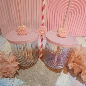 Puede incluir: Dos vasos de vidrio transparente con tapas y pajitas rosas, adornados con dijes de caritas sonrientes. Una pajita es rosa, la otra es a rayas rojas y blancas. Los vasos están sobre una superficie de madera con un fondo rosa y elementos decorativos.