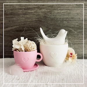 May include: A pink teacup with a heart design, filled with white flowers and a woven ball, sits next to a white teacup with a ceramic bird. The white teacup also has a heart design. Decorative elements include feathers and pearls.