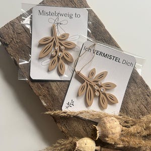 Könnte beinhalten: Zwei hölzerne Mistelzweig-Ornamente auf weißen Karten, verpackt in durchsichtigem Kunststoff. Die Ornamente sind hellbraun, mit einem Blatt- und Beeren-Design. Eine Karte lautet "Mistelzweig to" und die andere "Ich VERMISTEL Dich".