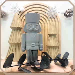 Puede incluir: Una figura gris de cascanueces con bigote y sombrero se encuentra frente a dos árboles de Navidad beige y un arco de madera. Copos de nieve decorativos y piñas adornan el fondo. Una ramita de hojas oscuras descansa en primer plano. El tema es Navidad.
