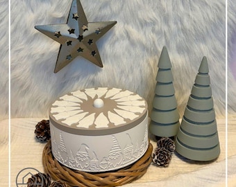 May include: A decorative holiday scene featuring a white and gray round box with a gnome design, two green cone-shaped trees, a metal star, and pinecones. The box sits on a woven base, creating a festive display.
