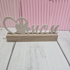 Puede incluir: Un letrero de madera gris claro con la palabra «MOM», un corazón y un diseño de huella de pata. Se apoya sobre una base rectangular de madera. El fondo presenta una pared rosa arqueada y una superficie de madera blanca.