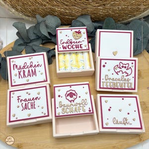 Könnte beinhalten: Eine Sammlung kleiner, rechteckiger Boxen mit weißen Deckeln und rosafarbenen Basen. Jede Box hat ein anderes Design und deutschen Text, darunter "Erdbeer-Woche" und "Baumwoll Schafe". Einige Boxen sind geöffnet und zeigen den Inhalt.